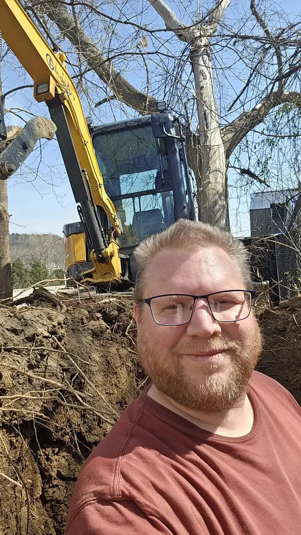Nick Strupp smiling beside an excavator at a dig site in the woods.
