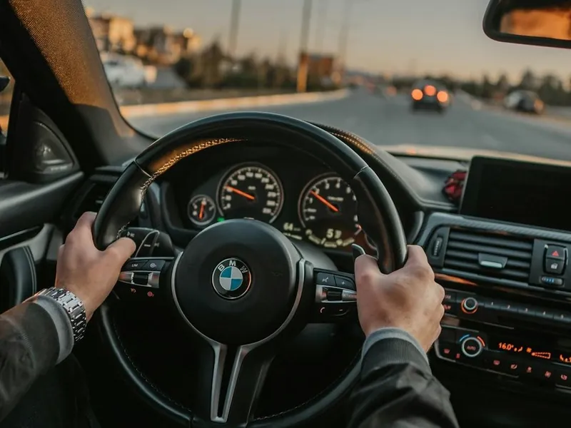 A driver’s hands on a steering wheel, looking down a city road at sunset.
